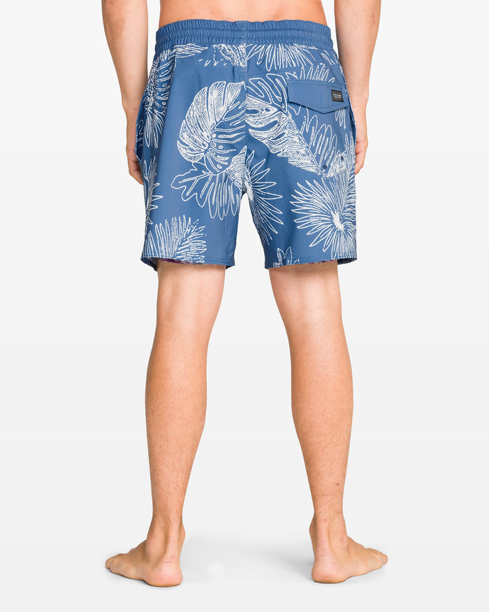 A person stands barefoot facing away, wearing Volcom Men's Lido Print 16" Trunk swim shorts with a white tropical leaf and flower print, featuring 4-way stretch, an elastic waistband, and a zippered back pocket.
