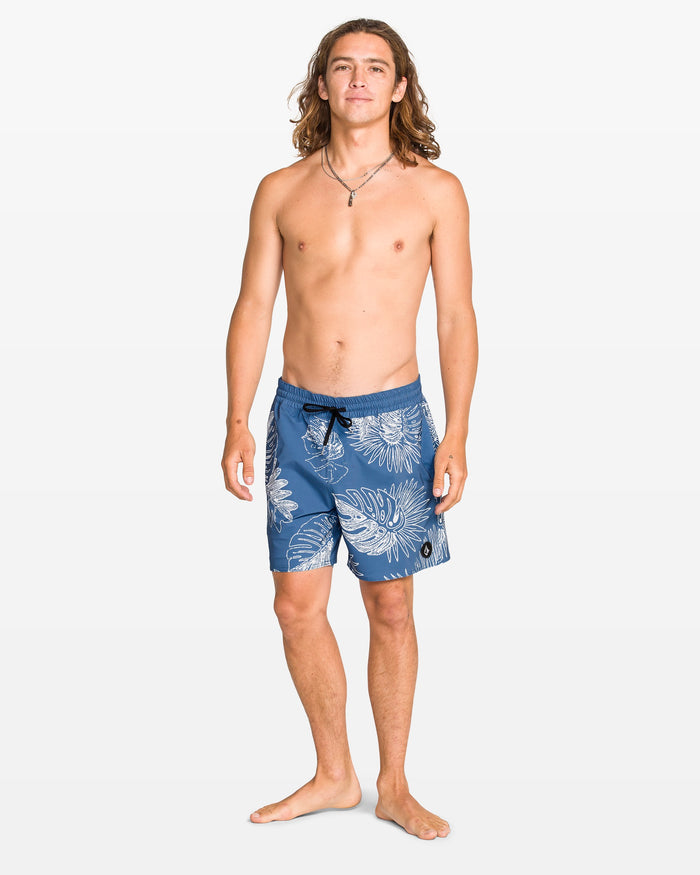 A young man with long hair stands barefoot on a white background, wearing Volcom Men's Lido Print 16" Trunk swim shorts. He is shirtless, smiling slightly, and has his arms relaxed at his sides.