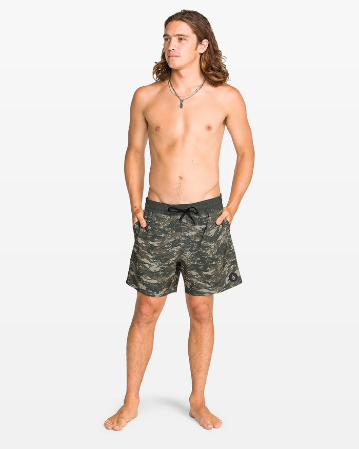 A barefoot man with long hair, wearing a necklace and Volcom Men's Lido Print Trunks in green camo with 4-way stretch, stands with his hands in his pockets against a plain white background.