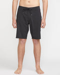 A person stands barefoot against a white background wearing Volcom Men's Lido Solid Mod 20 black knee-length swim trunks with a drawstring waist. Their hands are relaxed at their sides, and the upper body is partially out of frame.