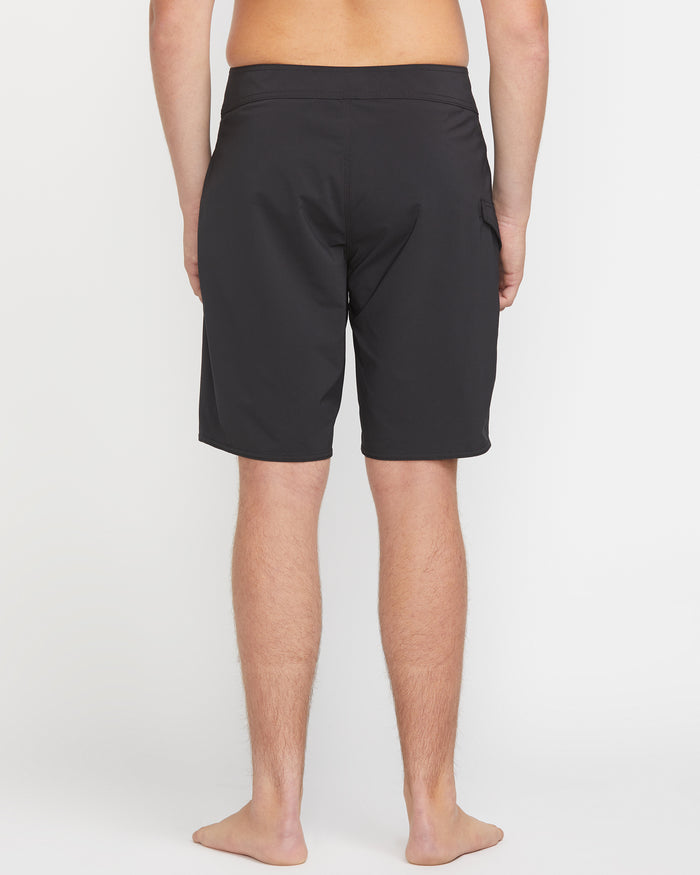 A person stands barefoot, facing away from the camera, wearing Volcom Men's Lido Solid Mod 20 black knee-length shorts against a plain white background.