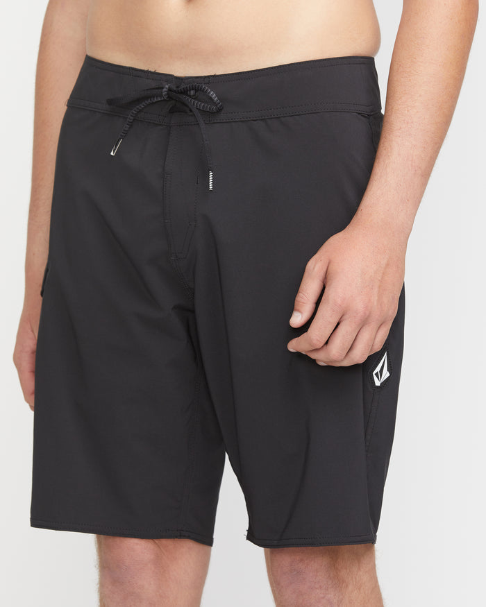 A man wears Volcom’s Men's Lido Solid Mod 20 black board shorts with a drawstring waist and small logo on the left leg, standing against a plain white background.