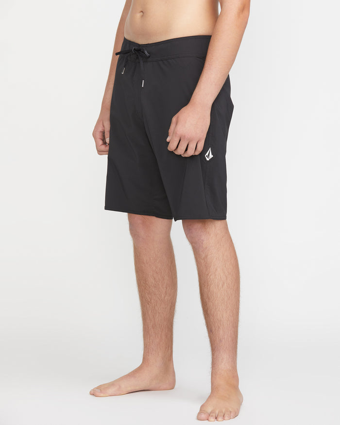 A man wears Volcom Men's Lido Solid Mod 20 black knee-length swim trunks with a drawstring waist and a small white logo on the left leg, standing barefoot against a plain white background.