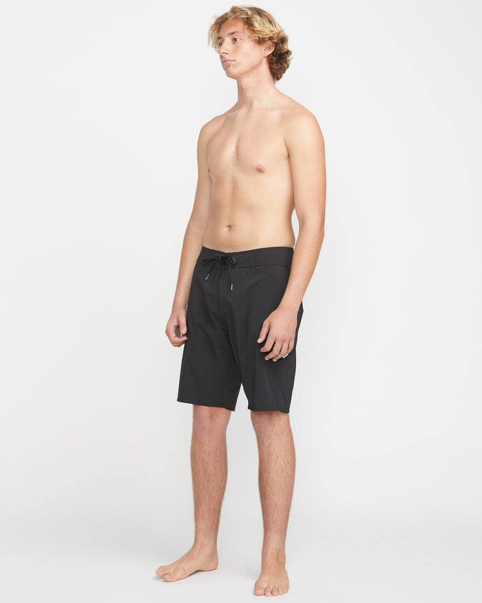 A young man with light skin and wavy blond hair stands barefoot against a plain white background, wearing Volcom Men's Lido Solid Mod 20 black knee-length swim trunks. He faces slightly left with his arms relaxed at his sides.