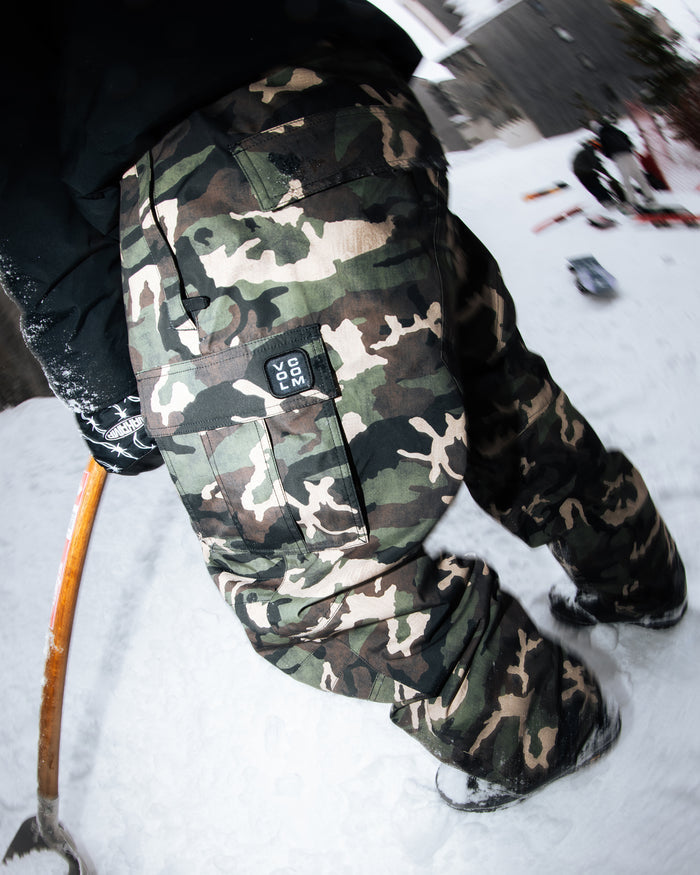 A person wearing Volcom Men’s Nwrk 20K Baggy Pants with a breathable lining stands on snow, holding a wooden-handled tool. The pants’ pocket features a visible ROC 200 patch. Nearby objects and snowy scenery appear in the background, captured with a fisheye lens effect.