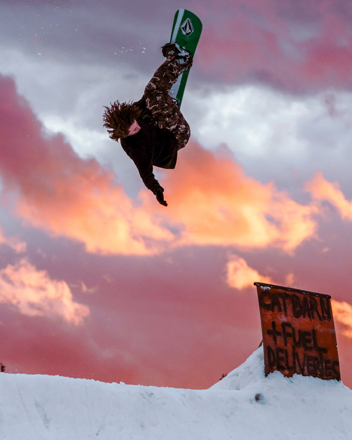A snowboarder wearing Volcom’s Men’s Nwrk 20K Baggy Pants performs a mid-air trick at sunset, with dramatic orange clouds overhead and a sign reading Cat Barn + Fuel Deliveries visible in the foreground.