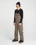 A man stands in a studio against a plain white background, facing slightly to the left, wearing a black beanie, long-sleeve black shirt, and Volcom Men’s Roan Bib with large pockets made from recycled stretch Oxford. He pairs the look with black shoes, showcasing both his style and the 15K protection of the Roan Bib.