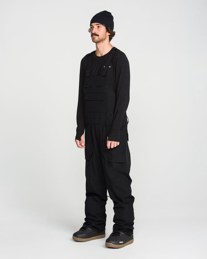 A person wearing a black beanie, long-sleeve shirt, utility vest, and the Volcom Men’s Roan Bib with adjustable suspenders stands against a plain white background, completing the look with gray sneakers.