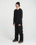 A person wearing a black beanie, long-sleeve shirt, utility vest, and the Volcom Men’s Roan Bib with adjustable suspenders stands against a plain white background, completing the look with gray sneakers.