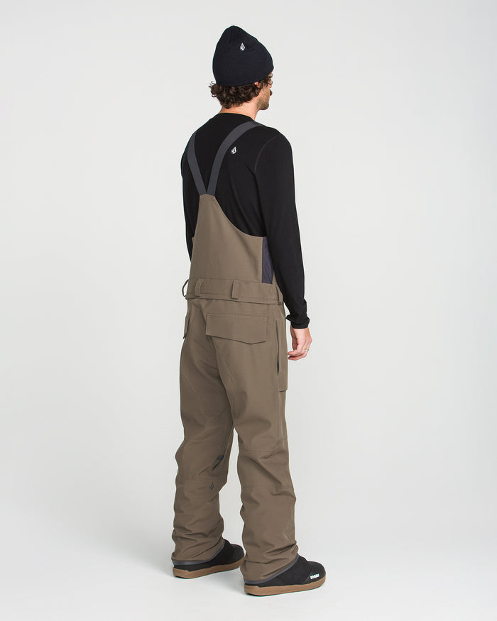 A person stands facing away on a plain white background, dressed in a black beanie, black long-sleeve shirt, and the Volcom Men’s Roan Bib Overall in khaki. Made from recycled stretch Oxford fabric, this bib showcases 15K protection.