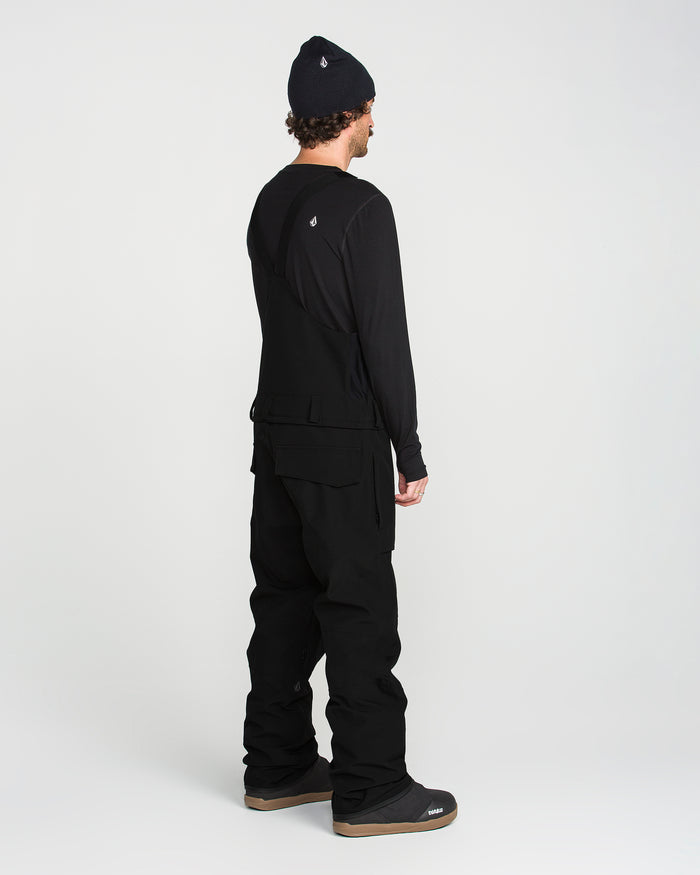 A person stands facing away, wearing a Volcom Men’s Roan Bib—a baggy recycled bib overall with adjustable suspenders—paired with a black beanie, long-sleeve shirt, and boots against a plain white background.