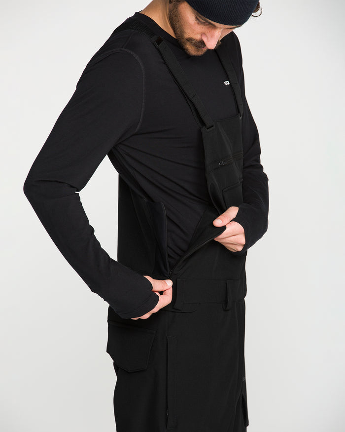 A person wearing a black long-sleeve shirt and the Men’s Roan Bib by Volcom, featuring adjustable suspenders, adjusts the side of their outfit against a plain white background.
