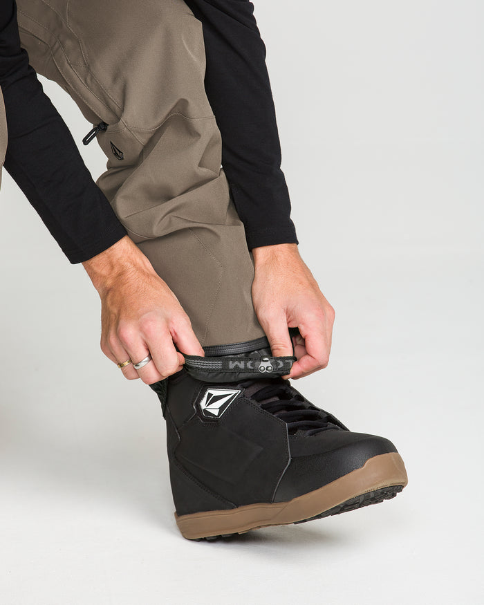 A person wearing Volcom Men’s Roan Bib Overalls in khaki, crafted from recycled stretch Oxford fabric, and a black long-sleeve shirt tightens the strap on a black snowboarding boot with a brown sole, ready to take on the slopes with 15K protection.