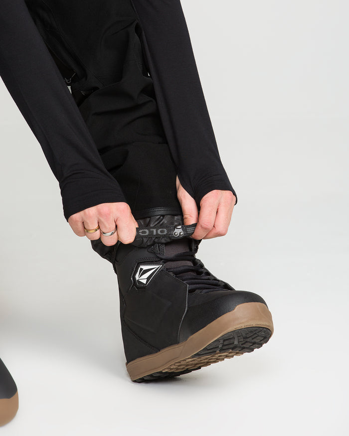 A person wearing Volcom Men’s Roan Bib black waterproof snow pants and a long-sleeve shirt is tightening the laces on a black, brown-soled snowboarding boot with both hands.