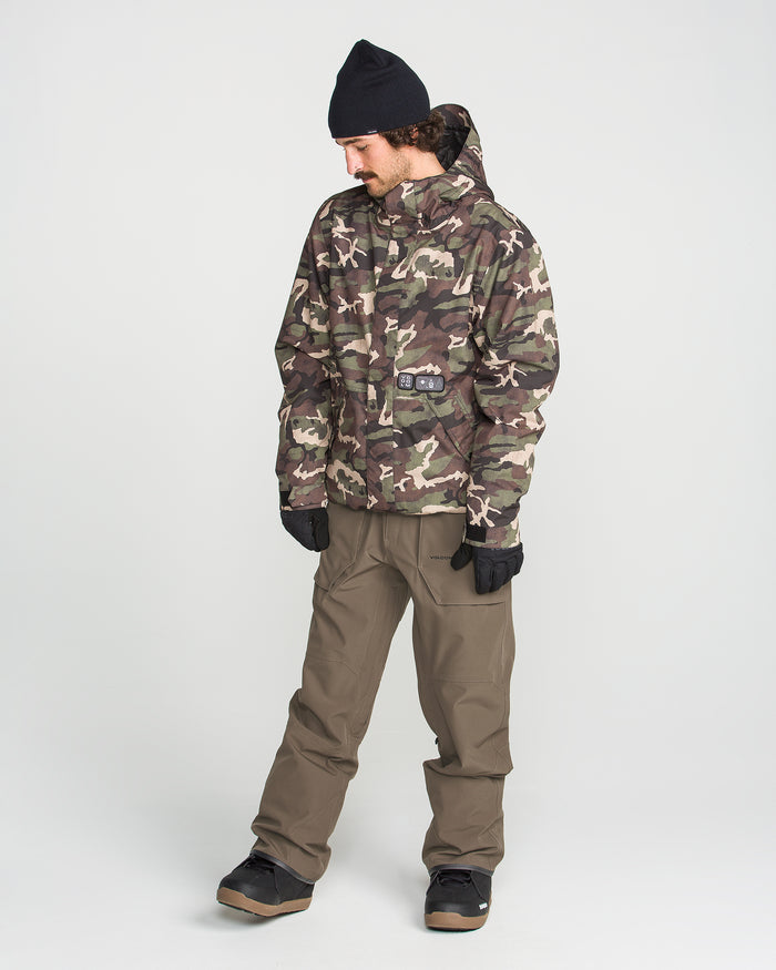 A person wearing the Volcom Men’s Roan Bib in camouflage print, along with a black beanie, brown pants, black gloves, and black shoes stands against a plain white background, looking down to their left.