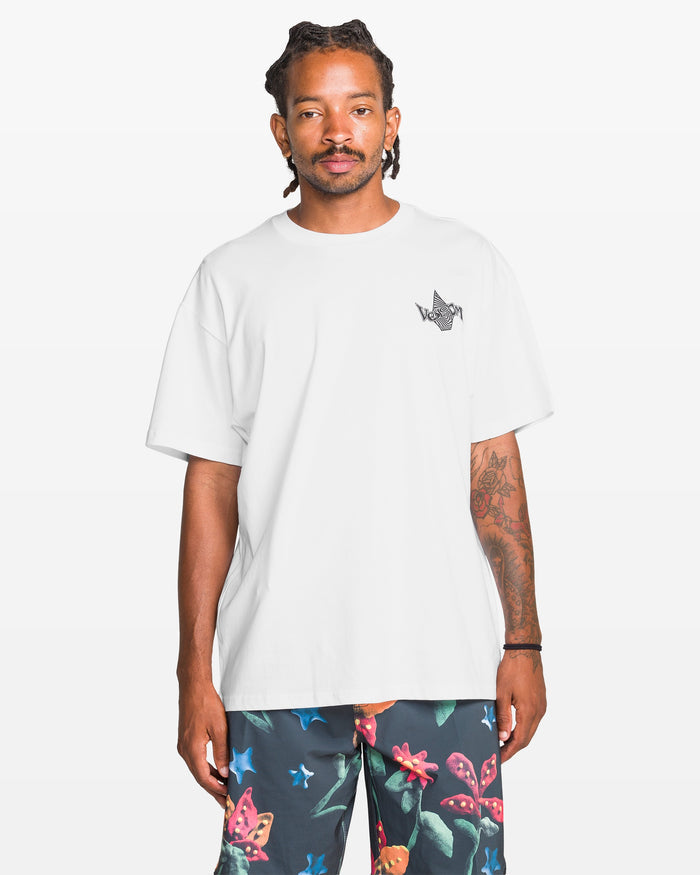 A man wearing the Volcom Men's Sea Legs Short Sleeve Tee with a small black chest graphic and dark floral shorts stands against a plain white background.