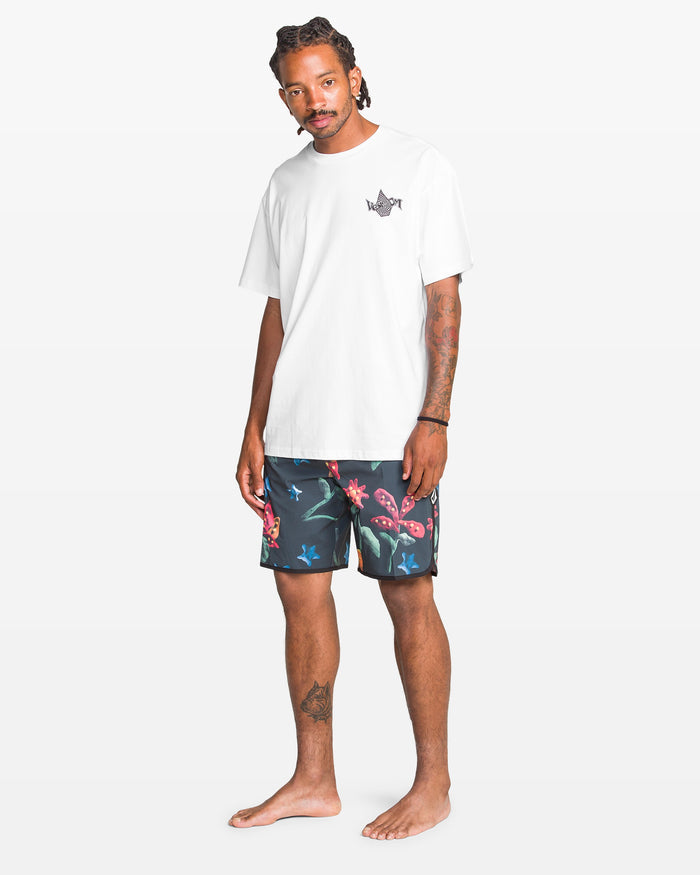 A man with braided hair and tattoos wears the Volcom Men's Sea Legs Short Sleeve Tee, a white heavyweight shirt with a small chest graphic, paired with dark floral shorts. He stands barefoot against a light, plain background.