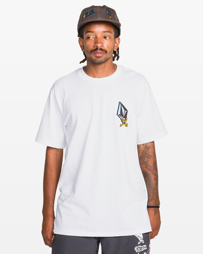 A man wearing the Volcom Men's Star Shields Featured Artist Exp Short Sleeve Tee, a 100% cotton white T-shirt with geometric graphic and cartoon figure, paired with dark shorts and a brown cap, stands against a plain white background.