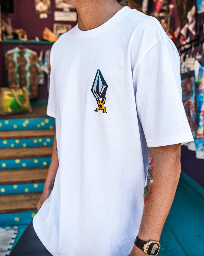 A person wearing the Volcom Men's Star Shields Featured Artist Exp Short Sleeve Tee, a 100% cotton white shirt with a geometric design and yellow Star Shields graphic on the chest, stands near blue stairs in a colorful store. Only their torso and arm are visible.