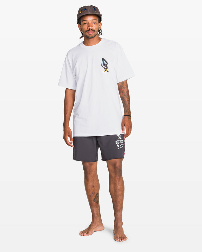 A man stands barefoot against a white backdrop, wearing the Volcom Men's Star Shields Featured Artist Exp Short Sleeve Tee in white, black printed shorts, and a black cap with blue and yellow designs.