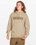 A person with long brown hair wears a beige Volcom Men's Stone Coast Pullover hoodie and brown pants, smiling against a plain white background. The hoodie features an orange and black Volcom logo and is made from brushed fleece with REPREVE recycled polyester.