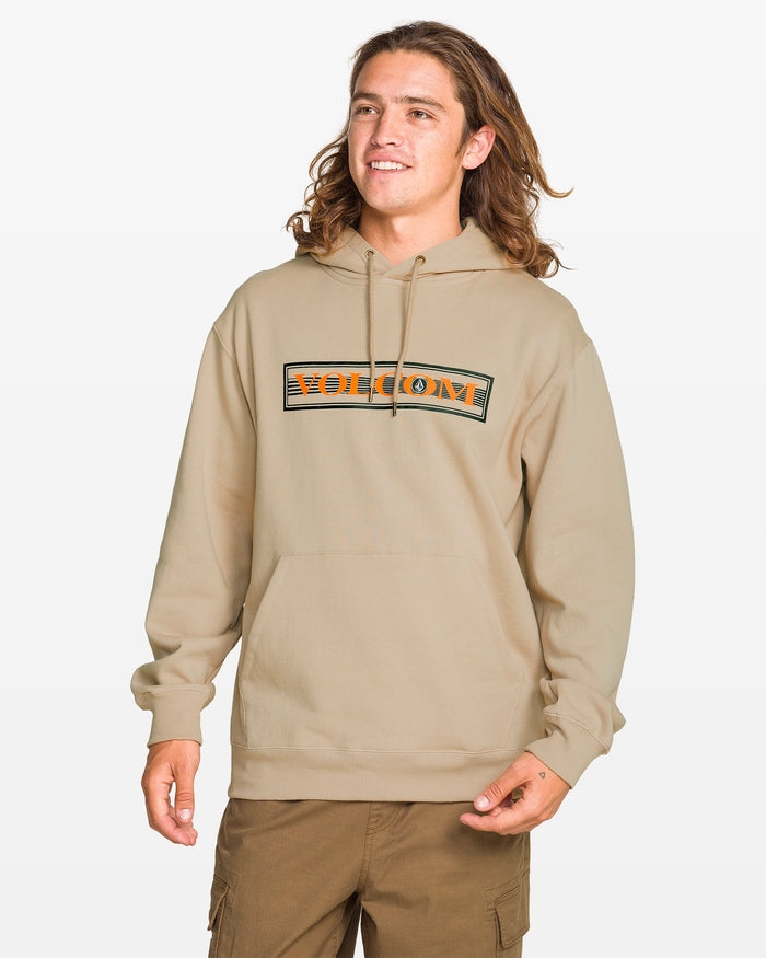 A person with long brown hair wears a beige Volcom Men's Stone Coast Pullover hoodie and brown pants, smiling against a plain white background. The hoodie features an orange and black Volcom logo and is made from brushed fleece with REPREVE recycled polyester.