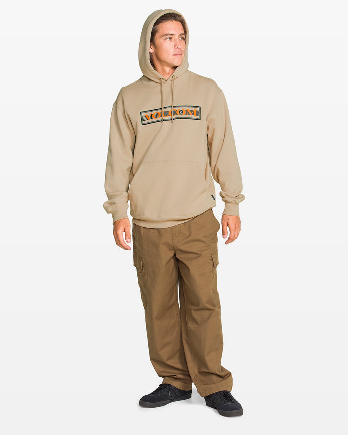 A man wears a tan Volcom Men's Stone Coast Pullover, brown cargo pants, and black shoes while standing slightly to the side against a plain white background.