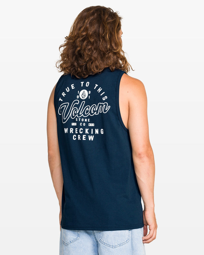 A person with long hair, seen from the back, wears a navy blue Volcom Men's Wrecking Tank in classic fit with "True To This, Volcom Stone Co, Wrecking Crew" printed in white on the back, paired with light blue jeans.