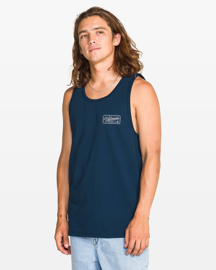 A man with long brown hair wears a navy blue Volcom Men's Wrecking Tank in a classic fit and light blue jeans, standing against a white background. The 100% cotton tank features a small white graphic on the left chest.