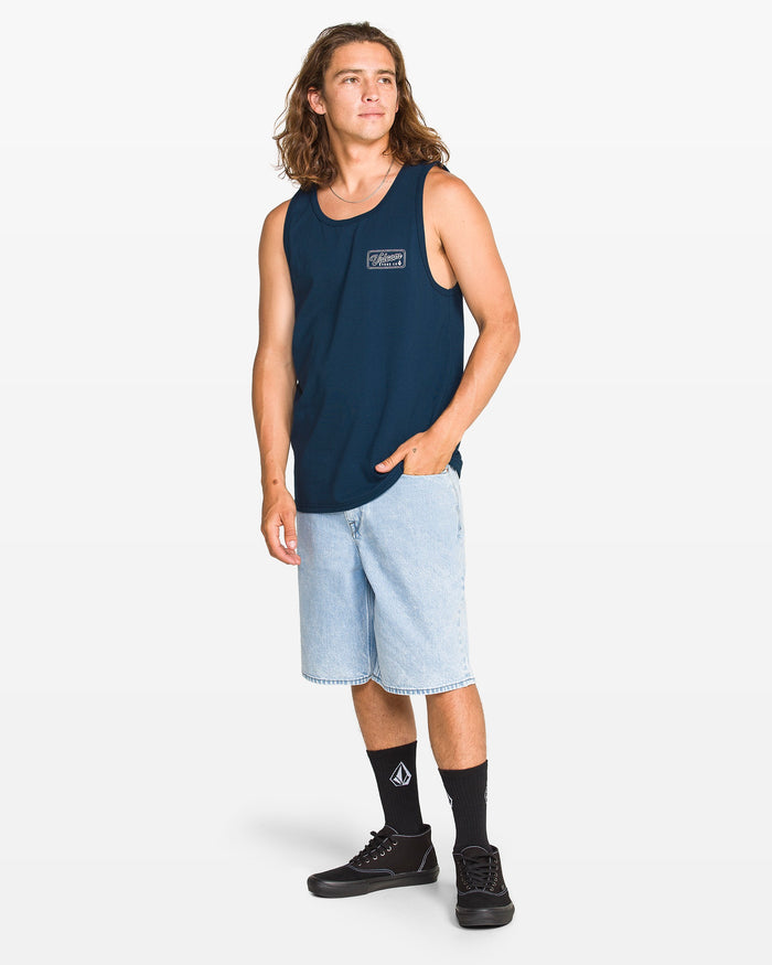 A young man with long brown hair models the Volcom Men's Wrecking Tank in navy, paired with light blue knee-length denim shorts, black logo socks, and black sneakers. He stands against a plain white background, looking slightly off to the side.