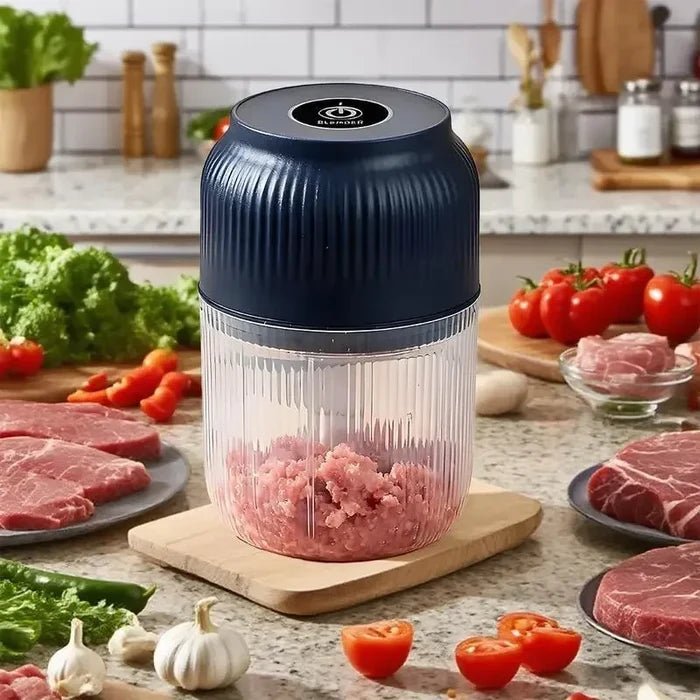 A countertop electric meat chopper with a dark navy blue lid and a clear ribbed container filled with ground meat, placed on a wooden cutting board in a kitchen setting with raw meat, tomatoes, garlic, and vegetables around it.