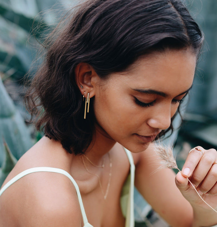 Mini Gold Bar Earring On Model Lifestyle. A close-up lifestyle shot of a woman wearing earrings outdoors.