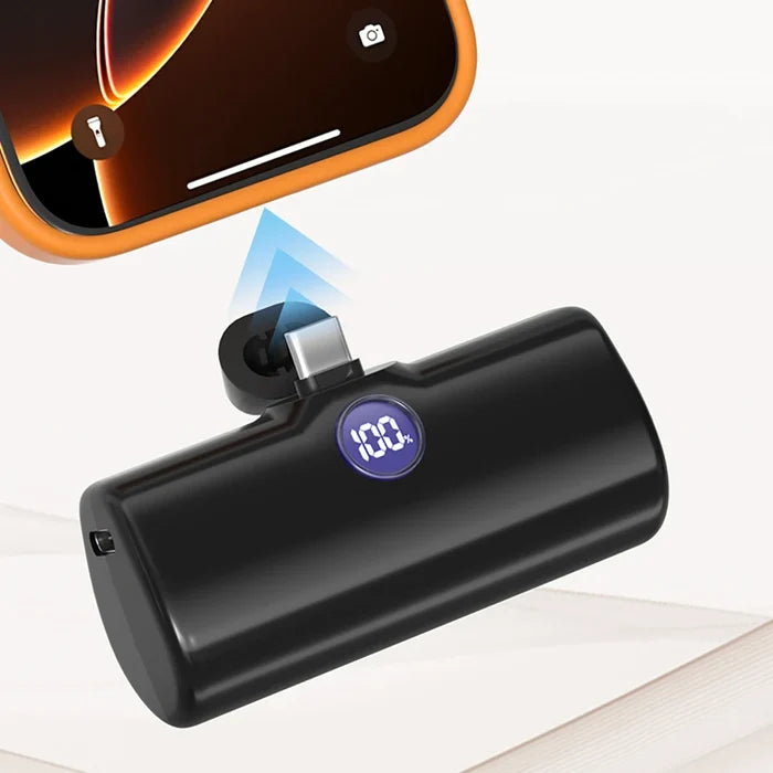 A black cylindrical portable power bank with a built-in USB-C port, shown charging a smartphone. The power bank has a small circular purple display reading 100% and is photographed on a clean white background with a phone and charging beam graphic.