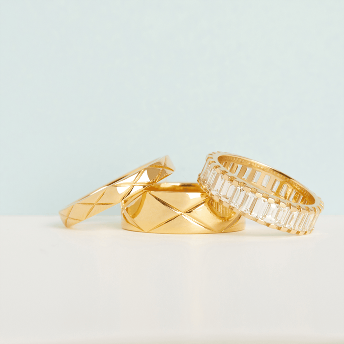 Lifestyle Model Close-up of three gold rings—two quilted bands and one diamond baguette band—resting on a white surface