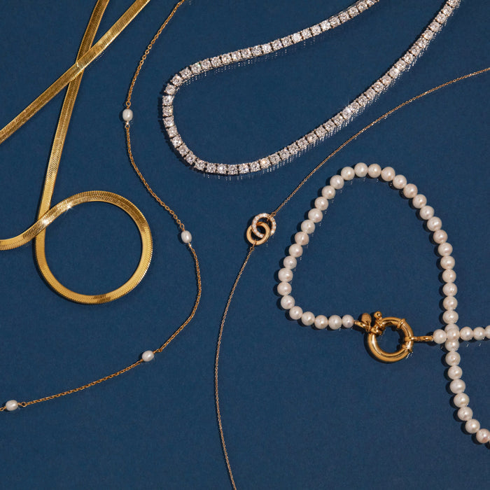 Flatlay Model Flatlay of five necklaces on a dark blue background, including a thick gold herringbone chain, a delicate chain with pearl stations, a diamond tennis necklace, a delicate chain with a circular gold pendant, and a pearl chain with a large round gold clasp.