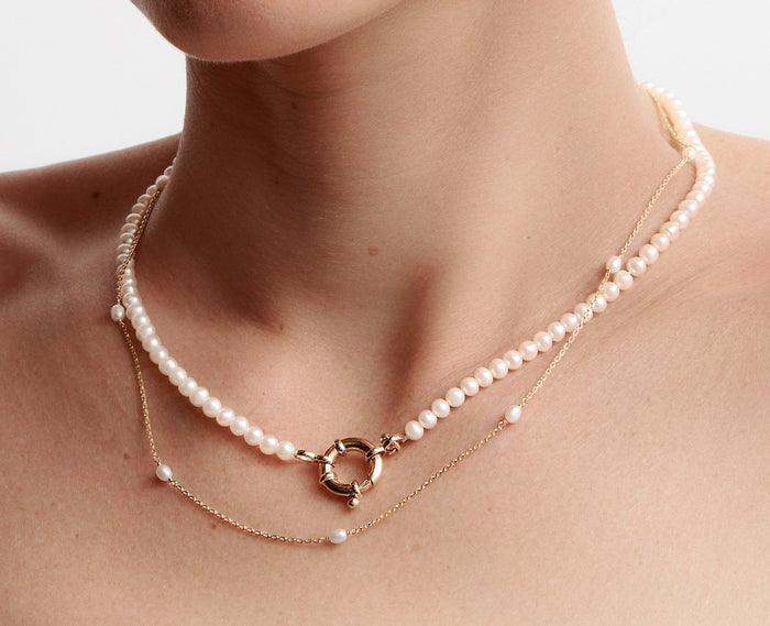 Lifestyle Model Close-up of a woman's neck wearing two layered pearl necklaces, including a full pearl strand with a large round gold clasp at the front and a longer, delicate gold chain with spaced-out pearl stations.