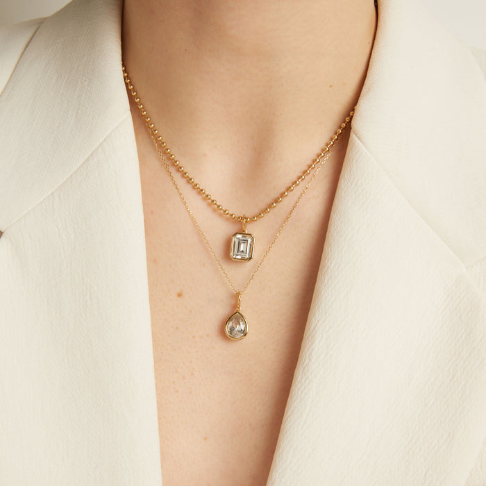 Model. Two gold necklaces with pendants on a person wearing a white outfit.
