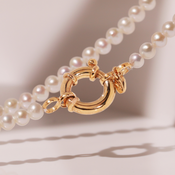 Model Close-up of a pearl necklace with a large round gold spring-ring clasp resting on a light beige surface.