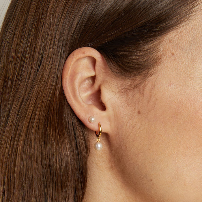Model. Close-up of an ear wearing a gold hoop earring with a pearl pendant.
