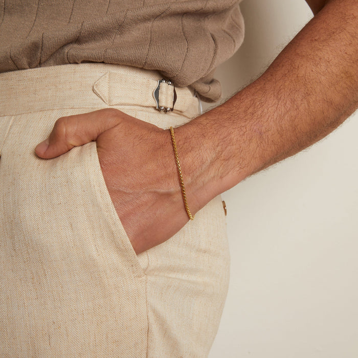 Model. Person wearing beige shirt and pants with a gold bracelet on a plain background