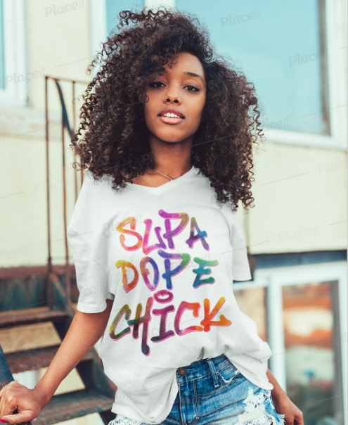 A woman with voluminous curly hair wearing a white graphic t-shirt that reads 'SUPA DOPE CHICK' in colorful bubble letters, paired with distressed blue jeans, posed outdoors against a blurred building background.