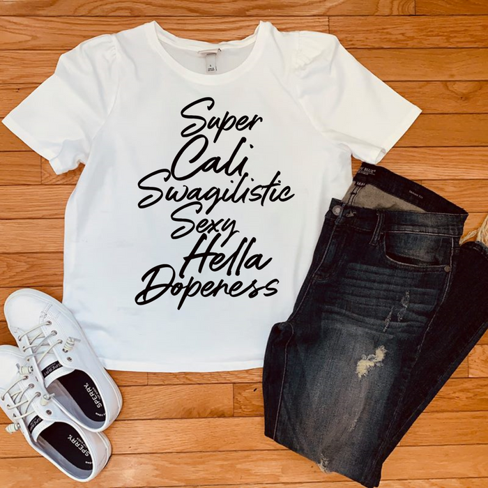 A white graphic t-shirt with black text reading 'Super Cali Swaguitistic Sexy Hella Dopeless' is styled in a flat lay on a wooden floor with dark distressed jeans and white sneakers.