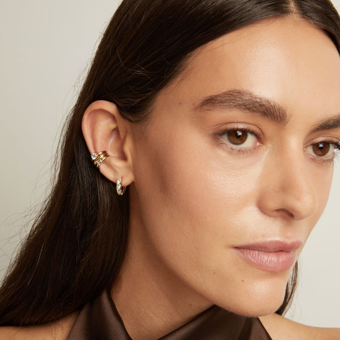 Model. Close up of woman wearing three gold ear cuffs and a huggie earring with white sapphires on neutral background.