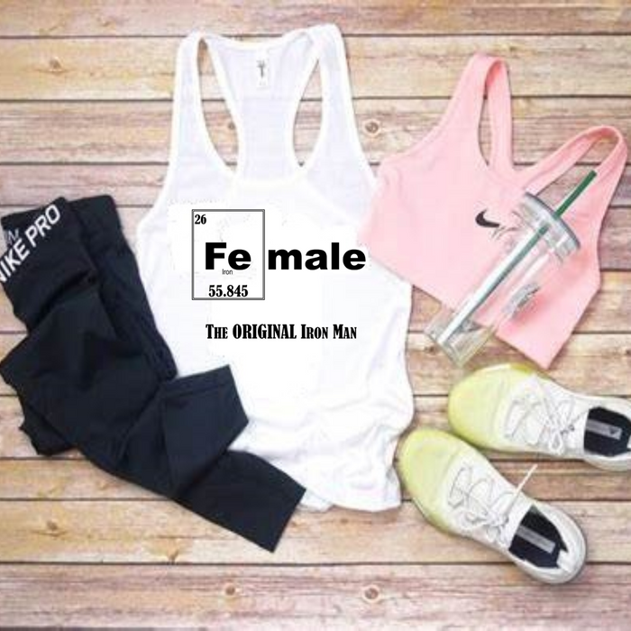 A flat-lay of athletic apparel on a wood plank background: a white racerback tank top with a periodic-table-style 'Fe male' graphic and text, a pink sports bra with a visible Nike swoosh, black leggings with white Nike text, a clear tumbler with a green straw, and white sneakers.