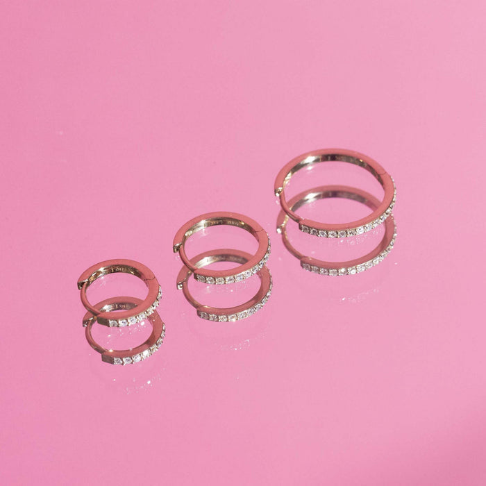 Flatlay Model Close-up of three pairs of gold and diamond hoop earrings on a bright pink background.