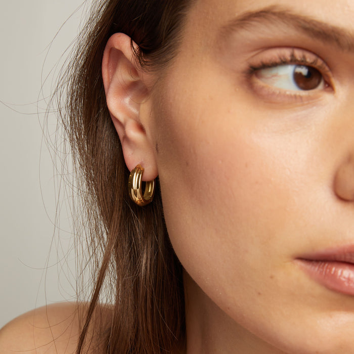 Model. Close up of a person wearing gold hoop earrings with a neutral background