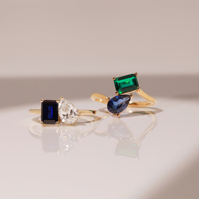 Model Close-up of two gold two-stone rings with different colored gemstones resting on a reflective white surface.