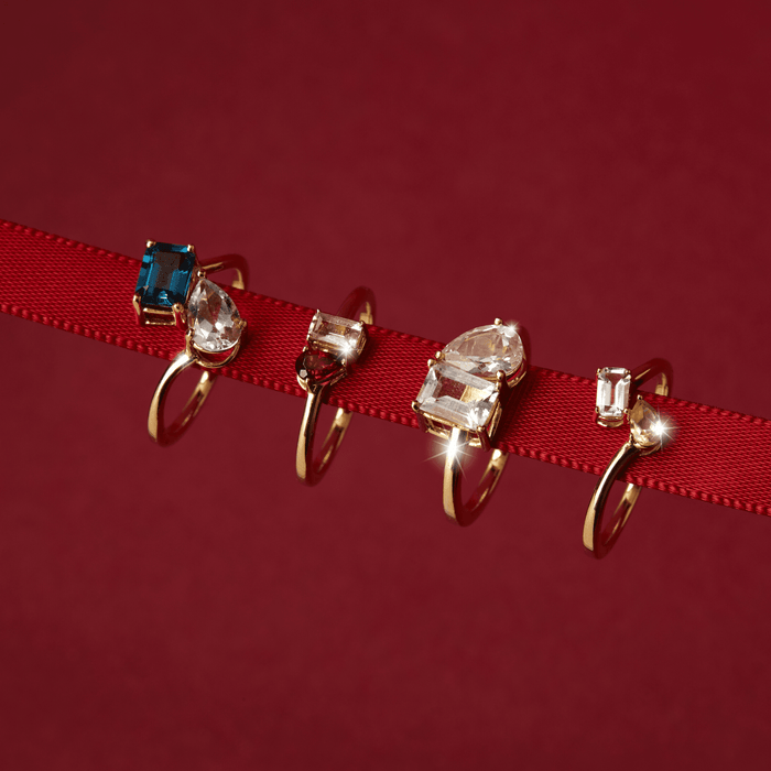 Flatlay Model Close-up of four gold two-stone rings with different colored gemstones resting on a red ribbon.