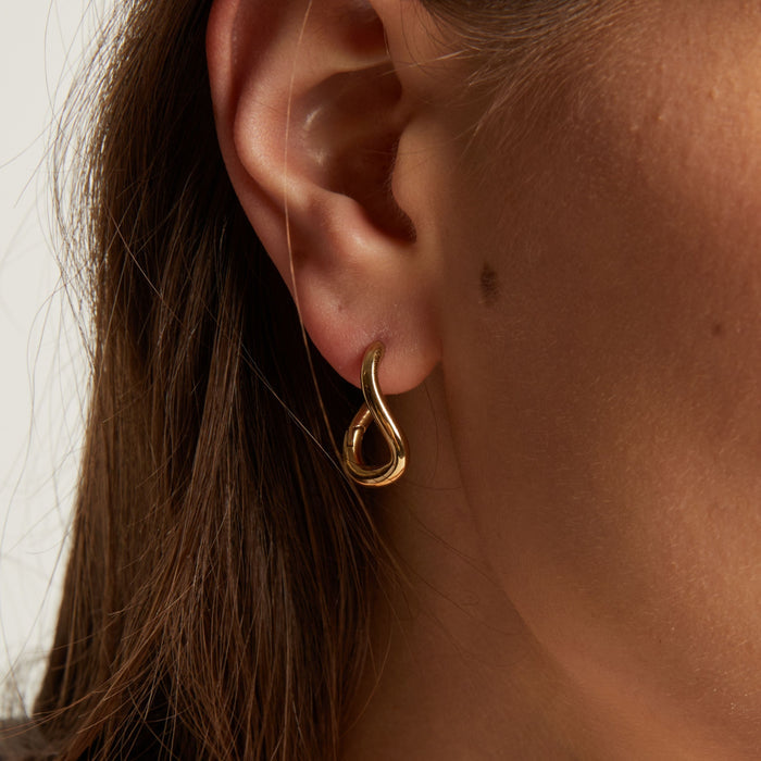 Model. Close up of a woman wearing gold twist earrings with a neutral background.