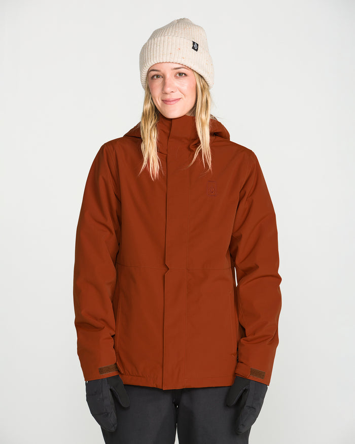 A person wearing a beige knit beanie, black gloves, and dark pants stands against a plain white background, smiling slightly at the camera while modeling the rust-colored Women’s Bolt Insulated Jacket by Volcom.